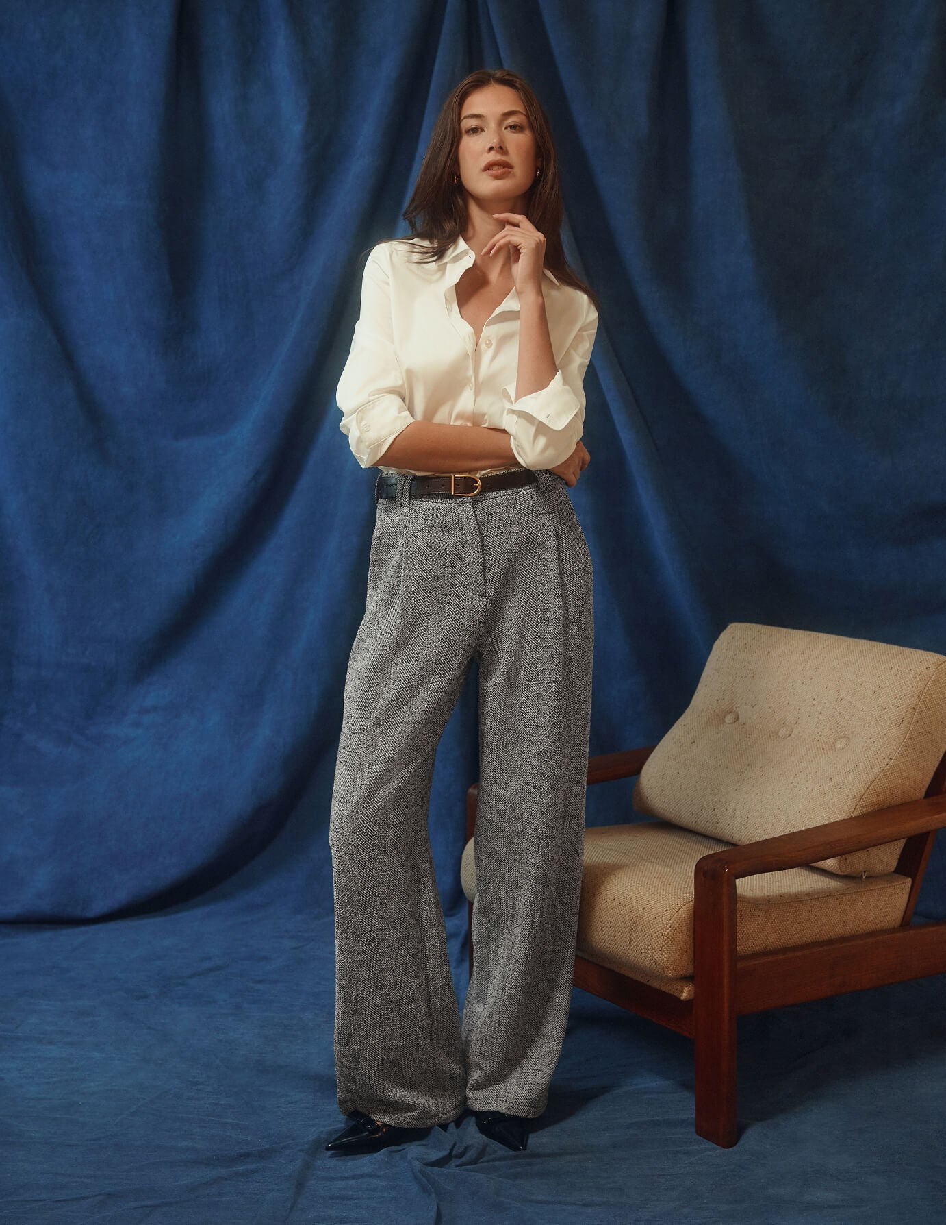 Eine Frau, die ein weißes Women’s Oxford Shirt White Orchid Slim und eine graue Hose trägt, steht vor einem blauen Stoffhintergrund.